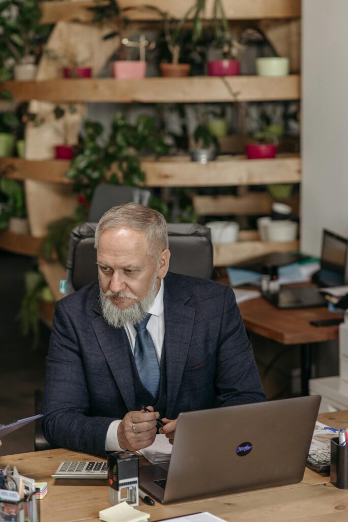Elderly professional in a formal suit working at a desk with a laptop in a plant-filled office.